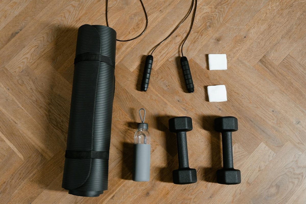 Flat lay of exercise essentials including dumbbells, yoga mat, and water bottle on wooden floor.