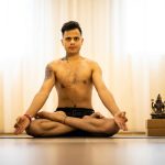 Man meditating in a lotus pose indoors with a statue beside him, promoting mindfulness.