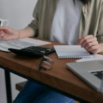 A woman manages finances at home, using a laptop and calculator on a wooden desk.