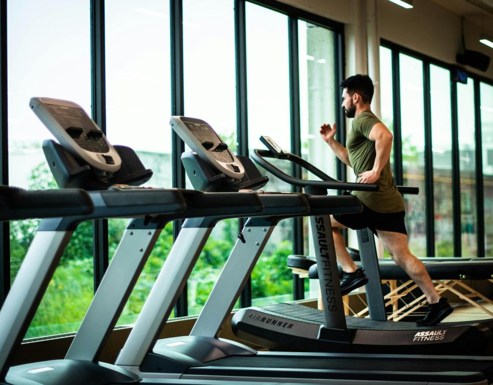 Young man workouts on treadmill in modern gym with large windows and natural light.