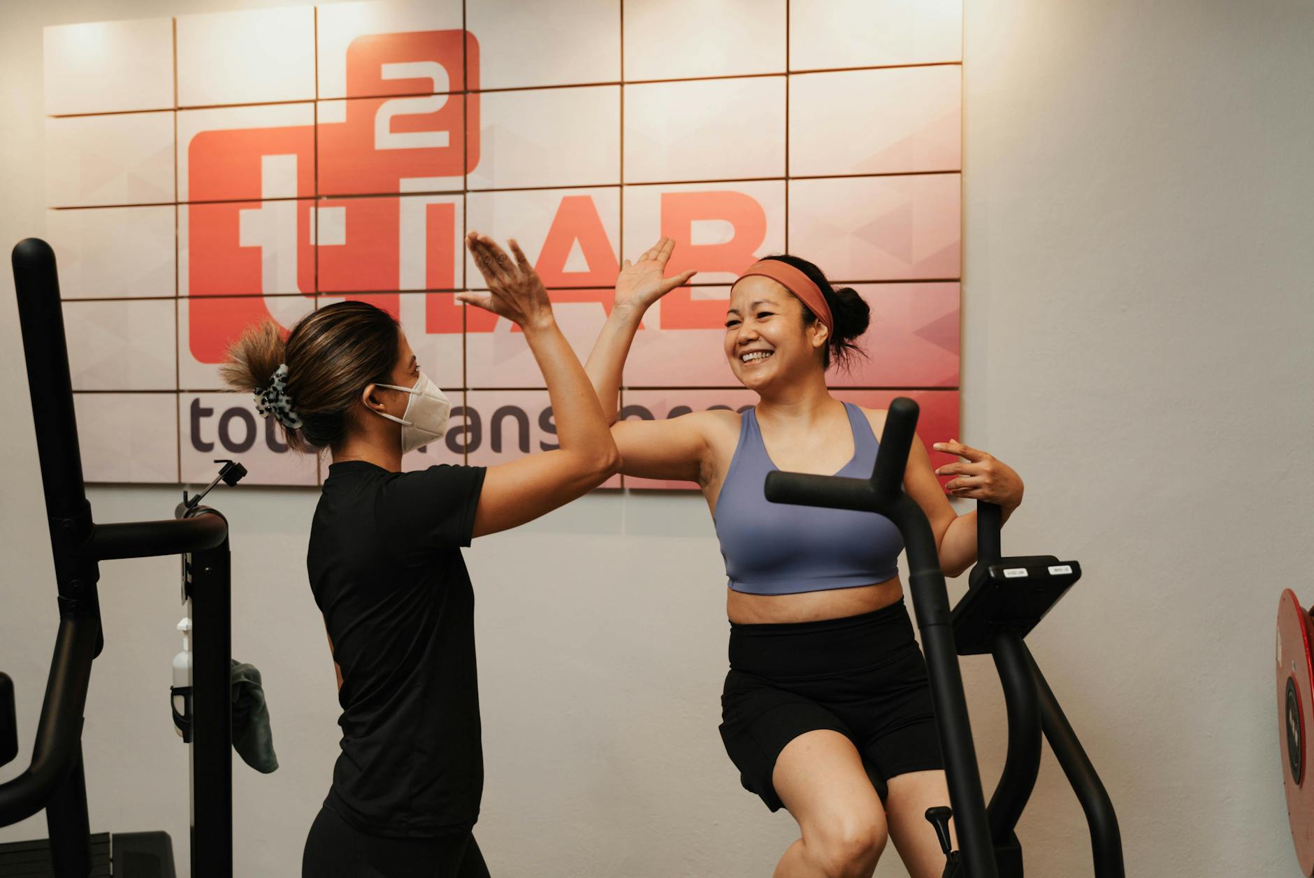 Two women engage in a joyful workout session, high-fiving at an indoor gym in Singapore.