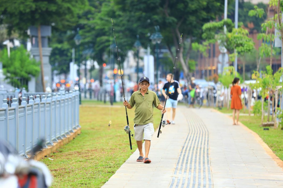 A man walks with fishing poles along a city park path, enjoying a sunny day outdoors.