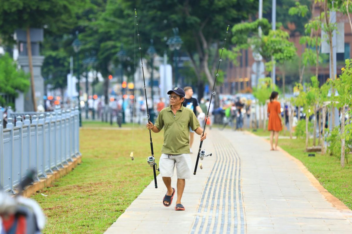 Man carrying fishing rods walks through a scenic city park during daylight.