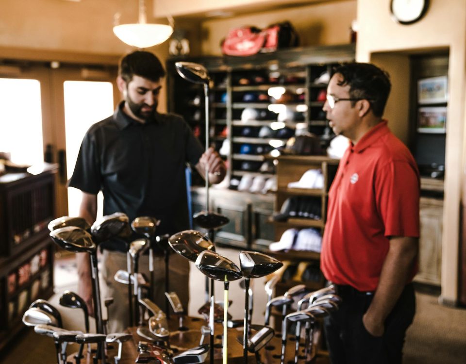 Two men browsing golf clubs in a sports equipment store, discussing options.