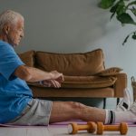 Elderly man in blue shirt exercising on a yoga mat indoors with dumbbells in view.