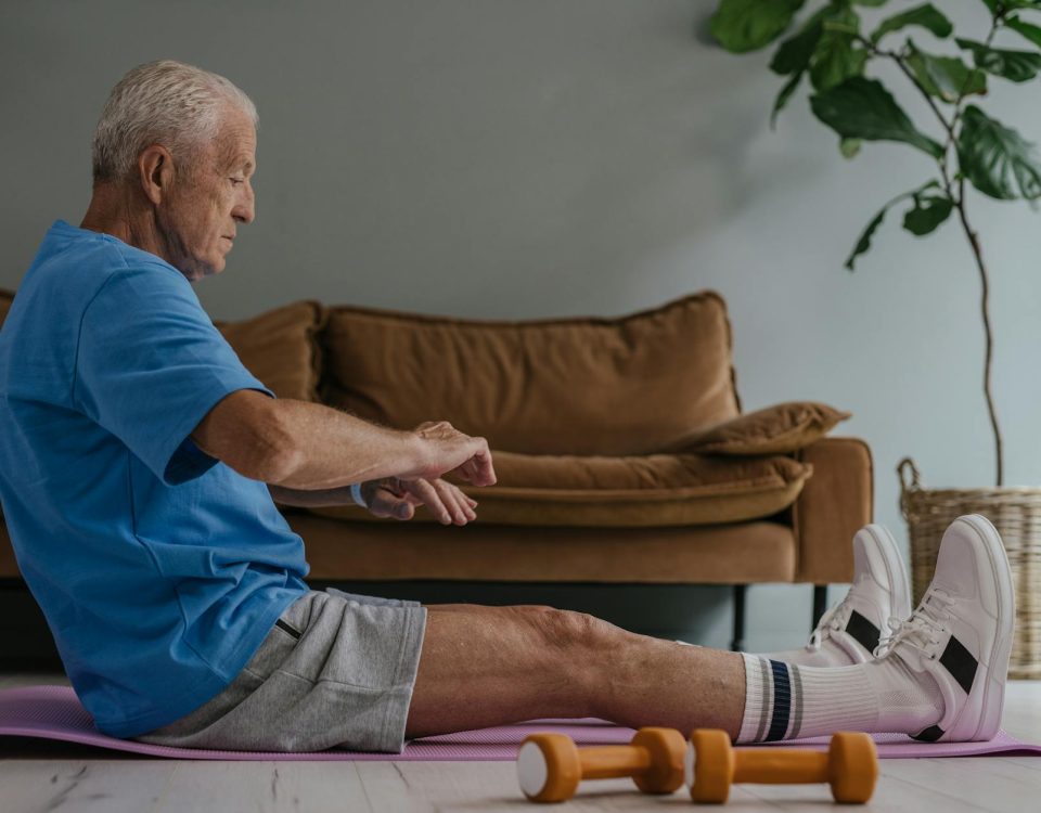 Elderly man in blue shirt exercising on a yoga mat indoors with dumbbells in view.