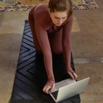 Woman in yoga attire kneeling on mat using laptop indoors, blending fitness with technology.