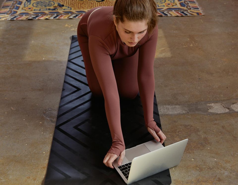 Woman in yoga attire kneeling on mat using laptop indoors, blending fitness with technology.