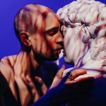 A man intimately engages with a sculpture, exploring themes of self-love and connection.