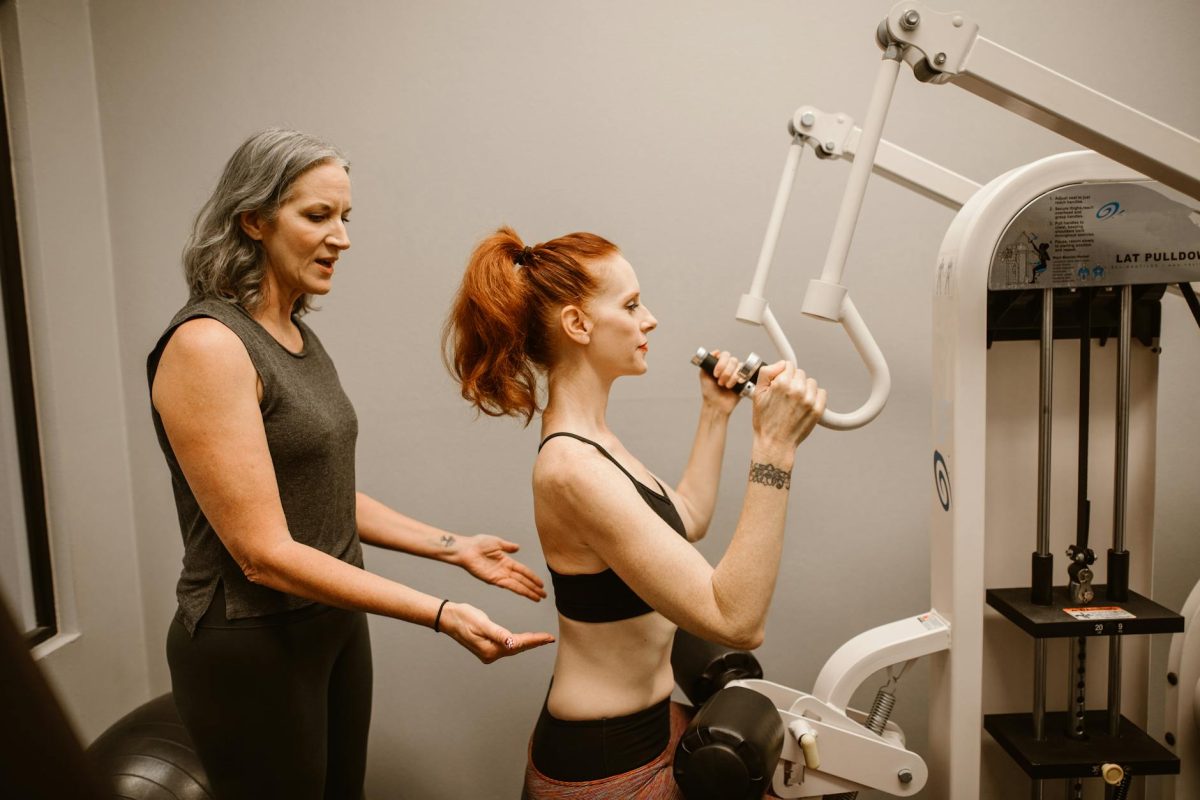 A woman works out on gym equipment with guidance from a trainer indoors.