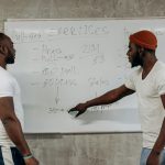 Two men discussing exercise routines on a whiteboard, planning gym workouts indoors.