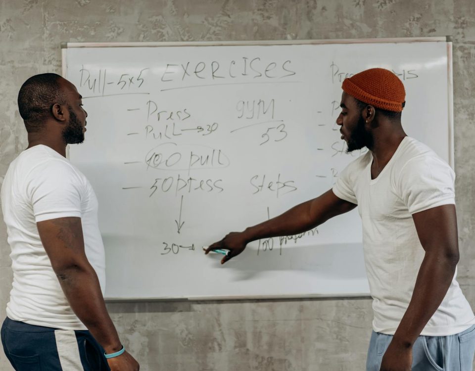Two men discussing exercise routines on a whiteboard, planning gym workouts indoors.