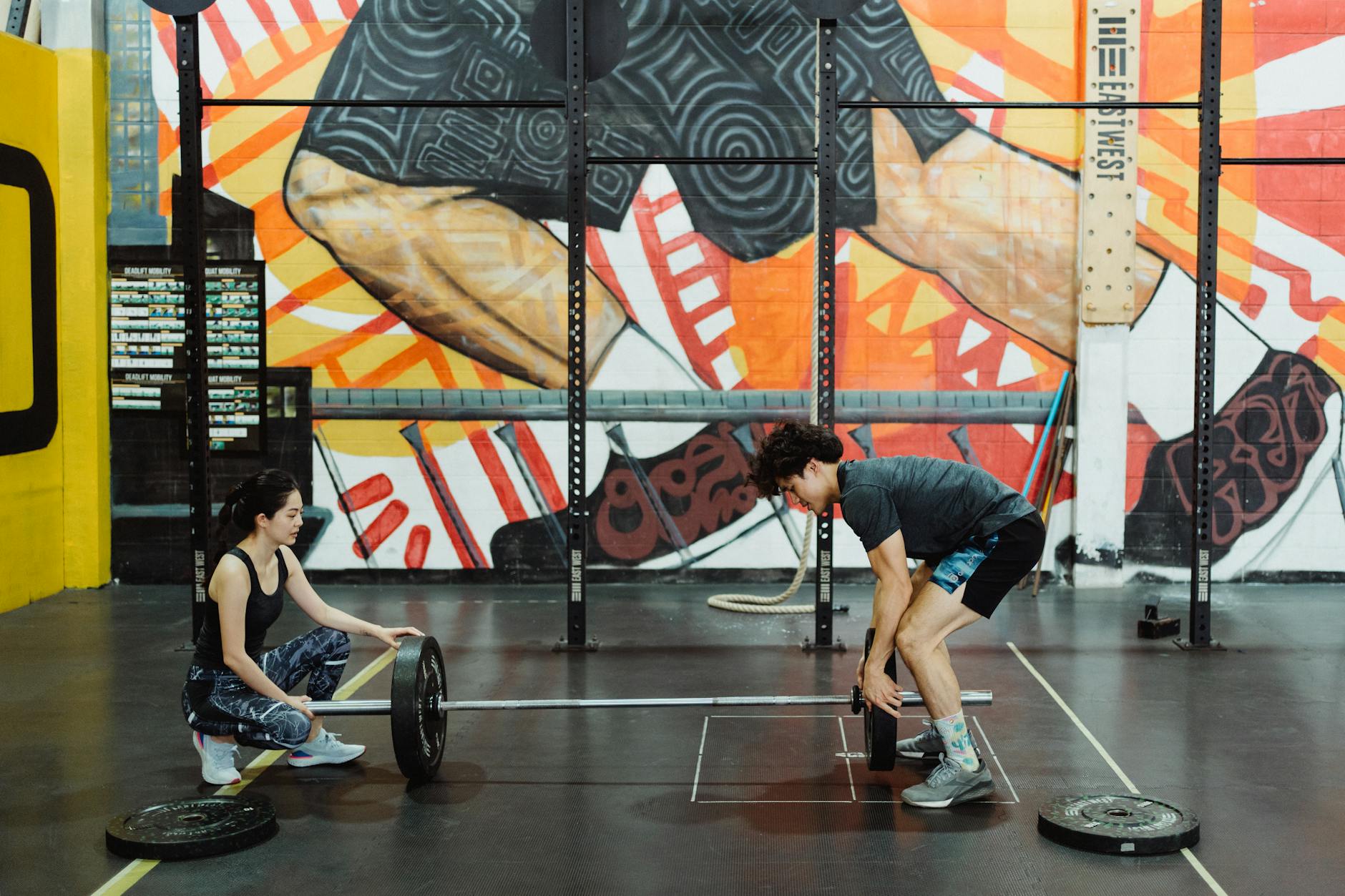 A man and woman engage in weightlifting training inside an art-filled urban gym.