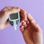 Hands holding a glucometer to test blood sugar levels on a purple background.