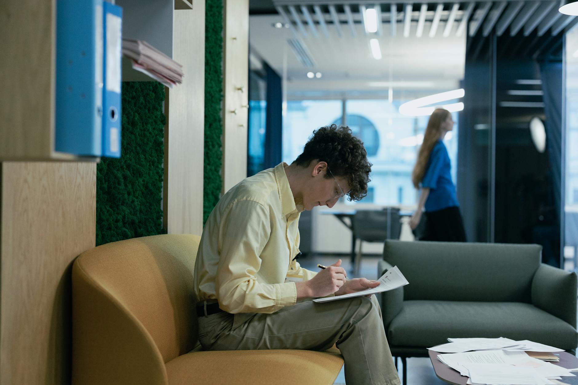 Office worker in yellow shirt writing notes on a couch, modern workspace with another person in the background.