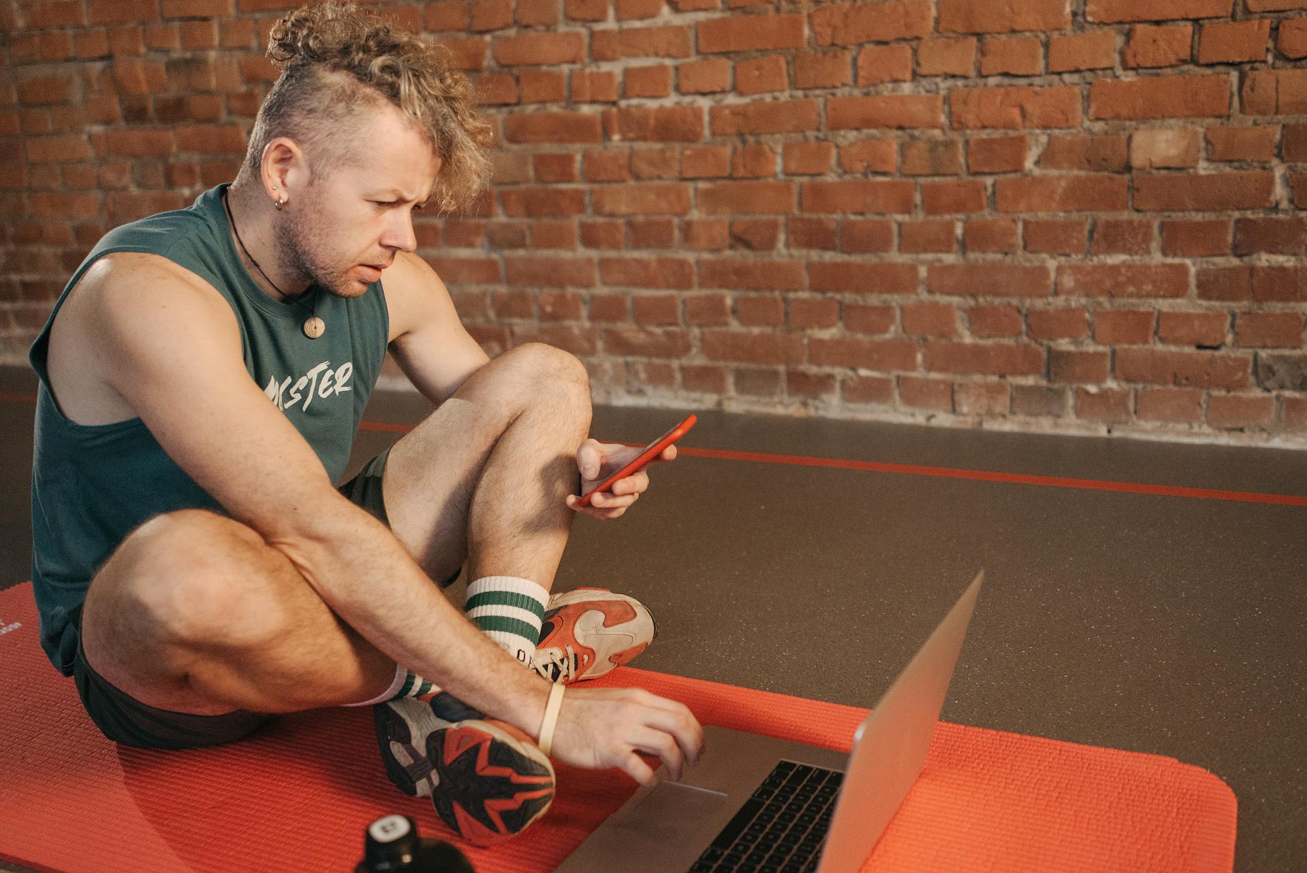 Man sitting on exercise mat using laptop and smartphone indoors.