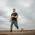 A muscular man running on sandy terrain under cloudy skies, promoting fitness and adventure.