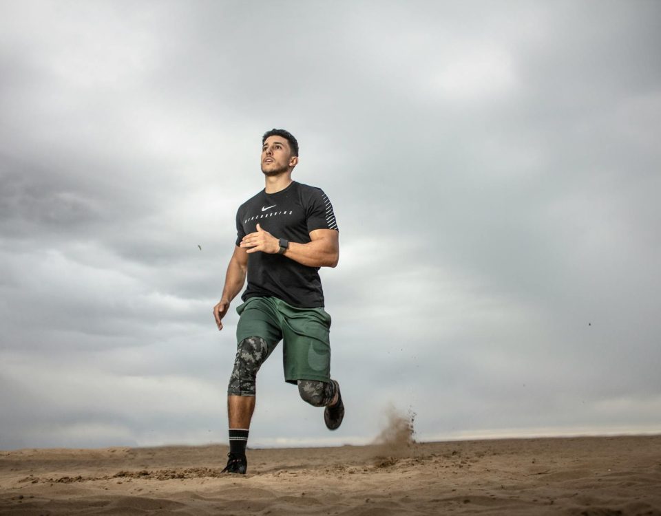 A muscular man running on sandy terrain under cloudy skies, promoting fitness and adventure.