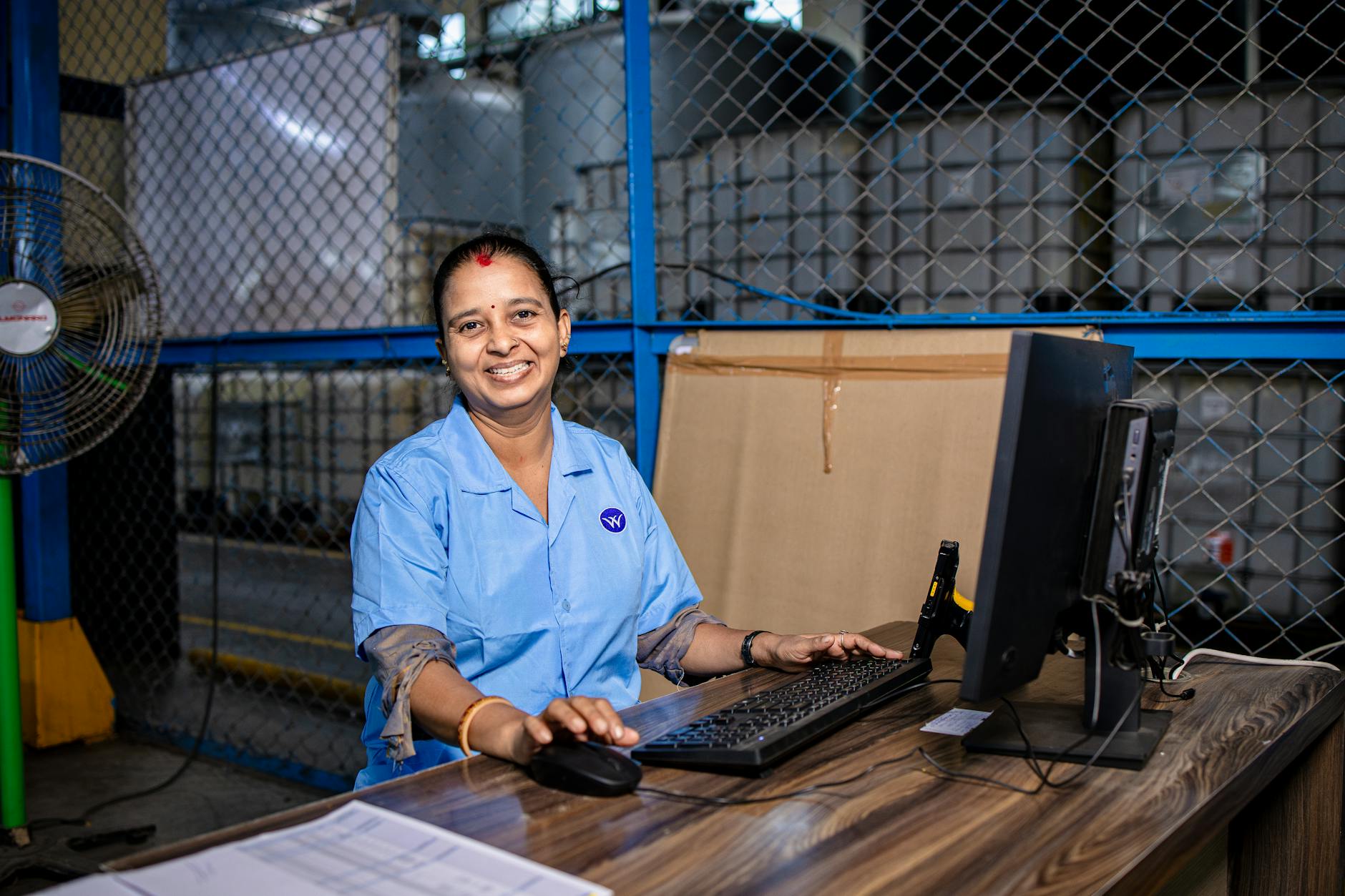 A cheerful female factory worker in blue uniform working at a computer in an industrial setting.