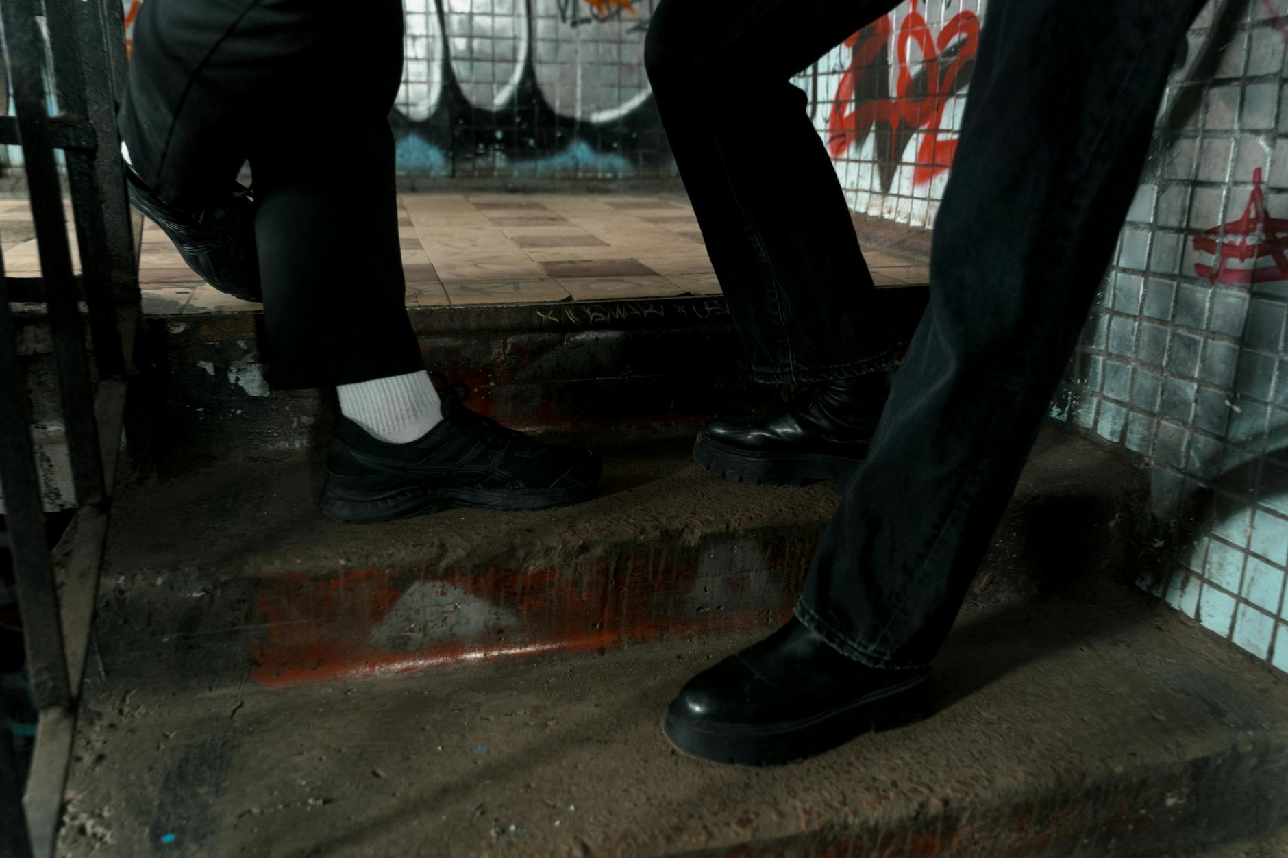 Close-up of two people wearing black shoes walking upstairs in an urban graffiti setting.