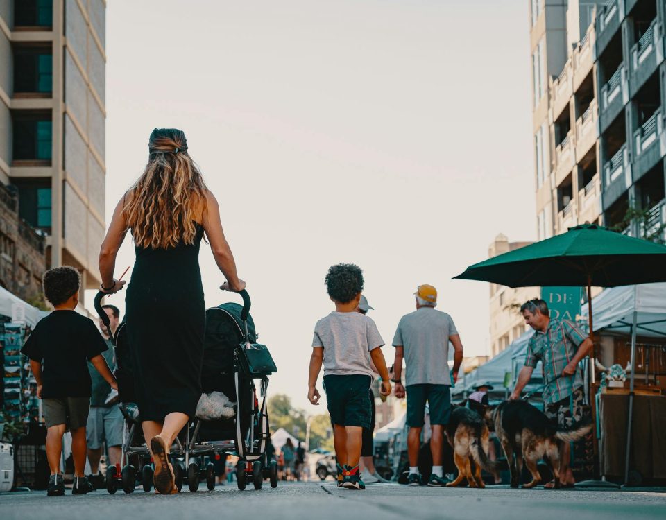 Family walking with stroller and dogs through lively outdoor market street on a sunny day.