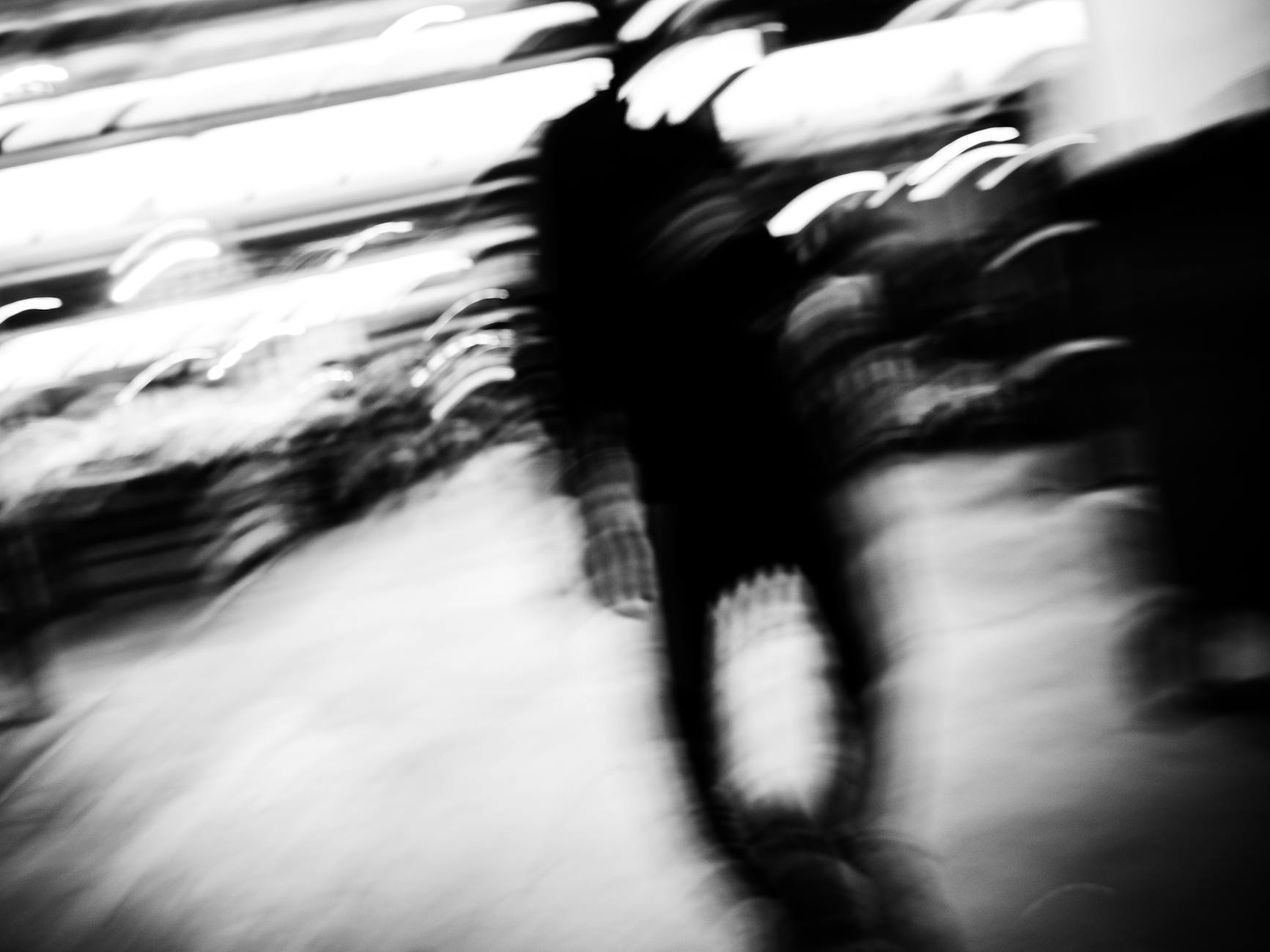 Blurred silhouette of a person walking in a busy urban setting, captured in black and white.