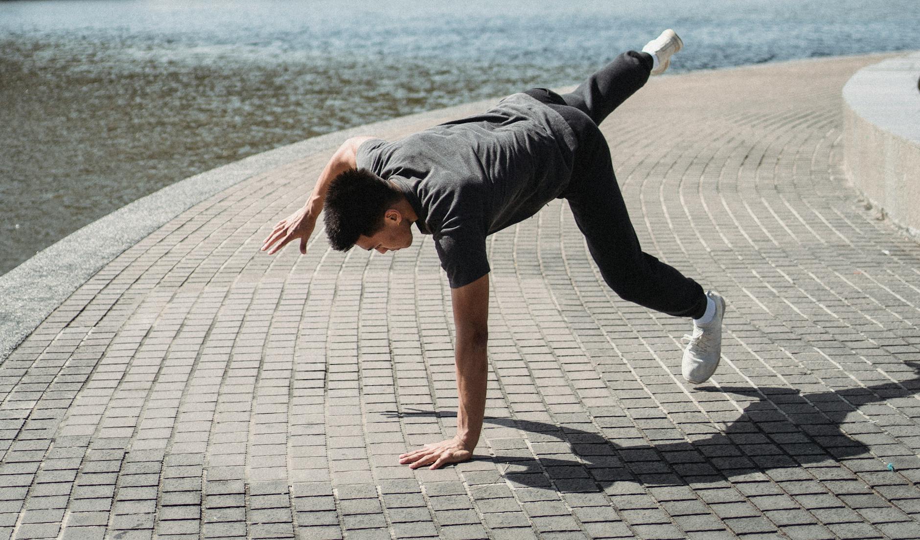 Anonymous young ethnic male athlete balancing on hand with raised leg near city river in summer